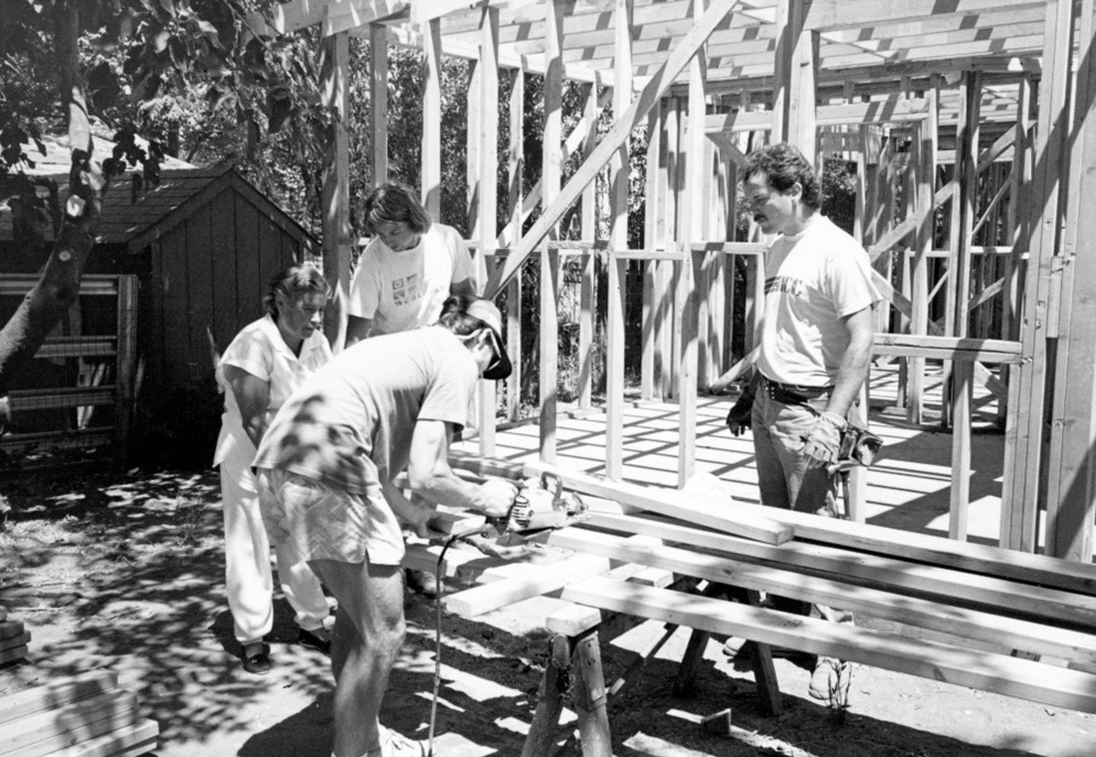 A black and white photo of volunteers working with Habitat for Humanity building the frame of a house. They are shown in front of the house, while one of the volunteers saws pieces of wood to add to the structure.