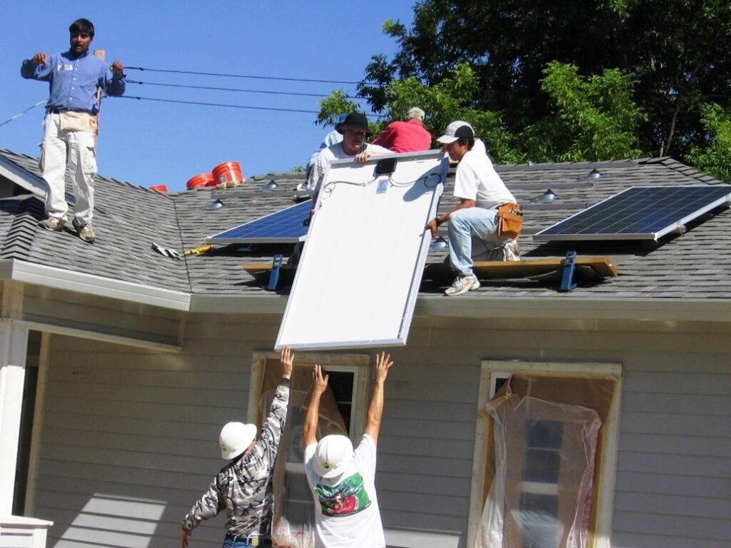 A photo of volunteers with Habitat for Humanity lifting solar panels onto the roof of a home to be installed.