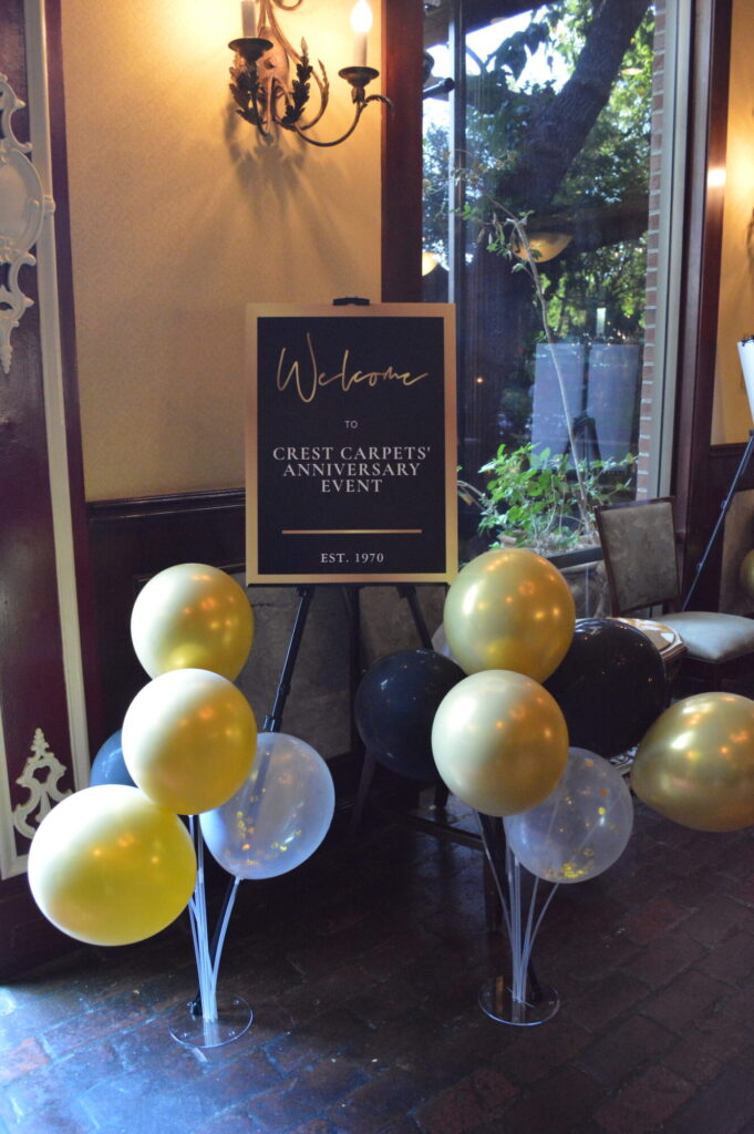A sign welcoming guests to the Crest Carpets Anniversary Event, surrounded by gold and black balloons.